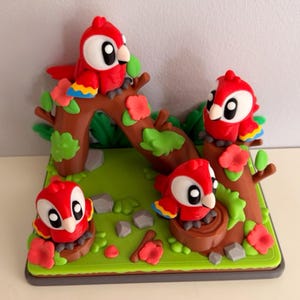 May include: A decorative figurine set featuring four red parrots with white faces and large black eyes, perched on brown tree branches and stumps. The scene includes green leaves, small red flowers, and grey stones on a square green base.
