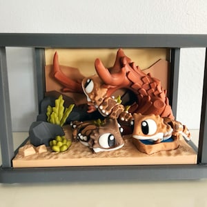 May include: A 3D printed diorama featuring three stylised lizard figures in a desert habitat. The scene includes a large brown horned lizard, two smaller lizards, green succulent plants, grey rocks, and a blue water dish, all enclosed in a grey frame.