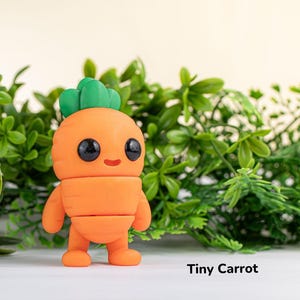 Può includere: Una piccola statuina a forma di carota arancione con una cima verde frondosa e occhi neri. La statuina ha un viso sorridente e si trova su una superficie bianca. "Tiny Carrot" è scritto in nero sotto la statuina.
