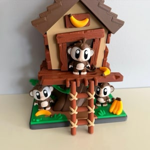 May include: A detailed figurine of a monkey treehouse, featuring three monkeys and bananas. The treehouse is brown with a ladder and a banana on the roof. The monkeys have large eyes and are holding bananas.