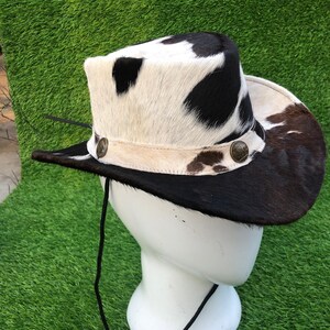 May include: A cowboy hat featuring a cowhide pattern with black and white patches. The hat includes a light-colored band adorned with two circular metallic buttons on the sides and a thin black chin strap hanging from the brim.