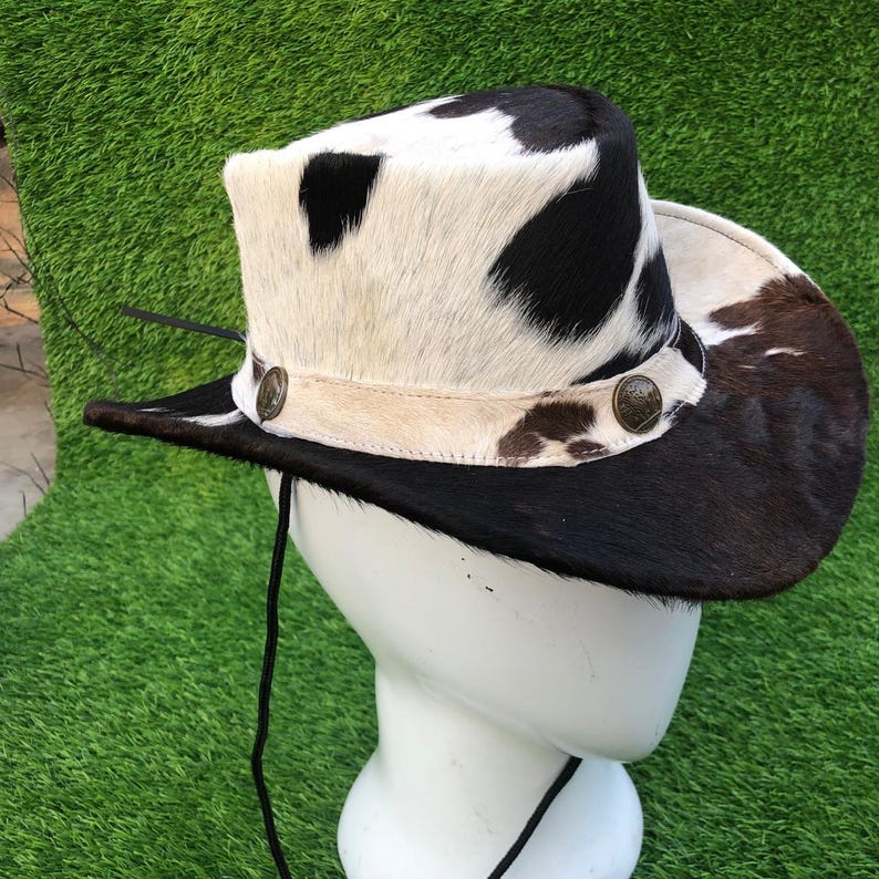 May include: A cowboy hat featuring a cowhide pattern with black and white patches. The hat includes a light-colored band adorned with two circular metallic buttons on the sides and a thin black chin strap hanging from the brim.
