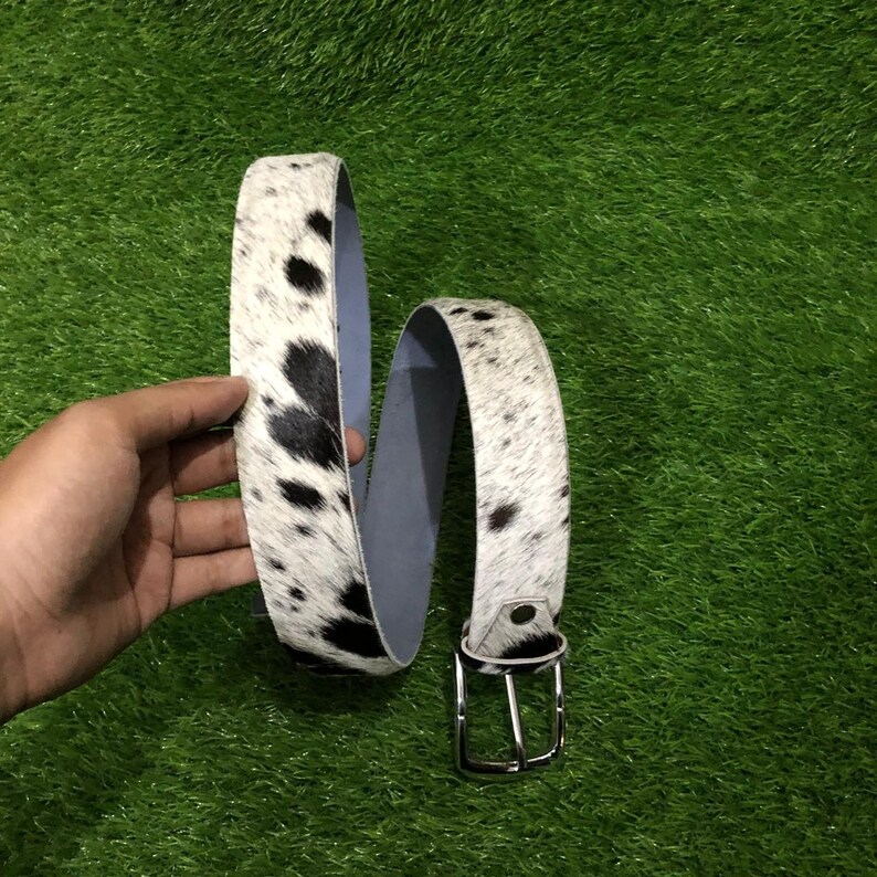 May include: A cowhide belt with a white background and black spots. The belt has a silver-colored buckle and a light blue interior. The belt is shown against a green background.