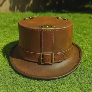 May include: A brown leather top hat with a circular brim and a cylindrical crown. The hat features a strap with a buckle and decorative metal accents. The hat is sitting on a green surface.