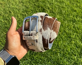 May include: A brown and white cowhide belt with a silver buckle. The belt is rolled up, displaying the natural pattern of the hide. The buckle is a classic, rectangular design. The belt is held against a background of green grass.
