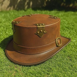 May include: A brown leather top hat with brass-colored hardware. The hat features a cylindrical crown, a wide brim, and decorative metal accents. The hat has a buckle closure on the band around the crown. The hat is sitting on green grass.