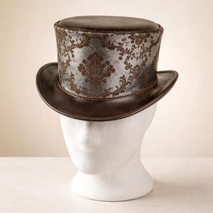 May include: A brown top hat with a decorative silver and brown patterned band. The hat has a rounded top and a wide brim. The hat is displayed on a white mannequin head.