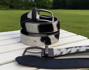 May include: Three leather belts with a black and white cowhide pattern are stacked on a white surface. One belt is unbuckled and lying flat. The buckles are a dark gunmetal colour. The background is a green field and trees.