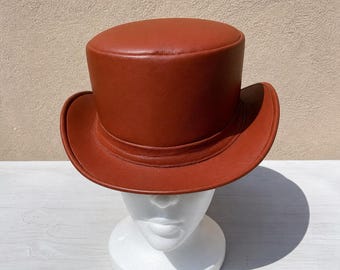 May include: A brown leather top hat with a rounded crown and a wide brim. The hat is displayed on a white mannequin head. The hat has a classic, formal design.