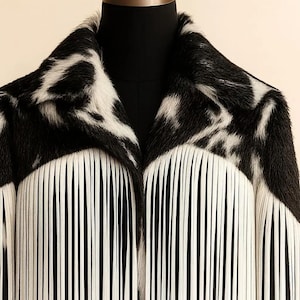 May include: A black and white faux fur coat with a striking pattern. The coat features a unique design with long, white fringe detailing along the front. The collar and upper portion of the coat showcase a distinctive black and white spotted pattern.