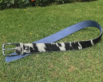 May include: A belt with a cowhide pattern in black and white, with a light blue leather backing. The belt has a silver-colored buckle and is lying on a bed of green grass.