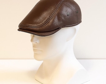 May include: A brown leather newsboy cap with a curved brim and two small buttons on the side. The cap is displayed on a white mannequin head, showcasing its shape and design. The cap has a classic, timeless style.