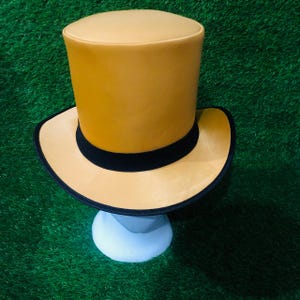Może przedstawiać: Musztardowy cylinder z czarną opaską i obszyciem ronda. Kapelusz jest prezentowany na białym stojaku na zielonym tle. Design kapelusza jest klasyczny i elegancki.
