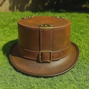 May include: A brown leather top hat with a circular brim and a cylindrical crown. The hat features a brown leather strap with a brass buckle and brass accents on the crown. The hat is sitting on green grass.