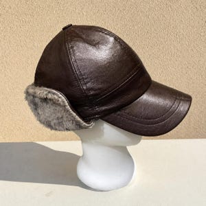 May include: A brown leather baseball cap with grey faux fur ear flaps. The cap has a curved peak and a button on top. The hat is displayed on a white mannequin head.