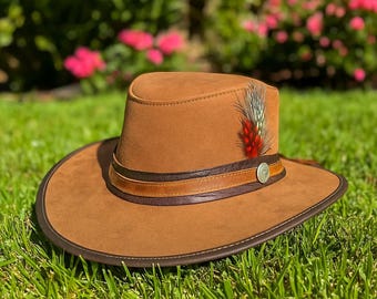 May include: A tan suede hat featuring a dark brown leather band, a lighter brown leather accent strip, and a small silver-toned circular emblem. A decorative feather with red and grey tones is tucked into the side of the hat band.