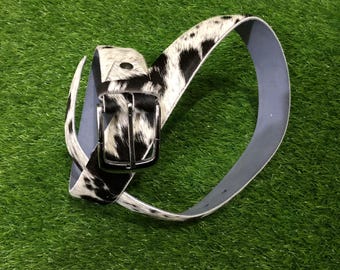 May include: A cowhide belt with a black and white spotted pattern. The belt has a silver-colored buckle and a light gray underside. The belt is displayed on a green surface.