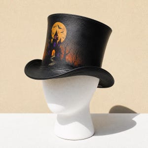 May include: A black top hat featuring a printed graphic of a haunted house silhouette against a large orange full moon. The design includes bare trees, a winding path, and small flying bats. The hat has a structured crown and a curved brim.