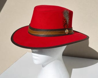 May include: A vibrant red hat with a wide brim and a flat top. The hat features a brown leather band with a gold button and a decorative feather. The brim is trimmed with a dark brown edge, creating a striking contrast. This hat is a stylish accessory.