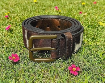 May include: A rolled-up brown leather belt with a brass buckle. The belt features a cowhide pattern with patches of brown and white. The belt is resting on a bed of green grass with small pink flowers scattered around.