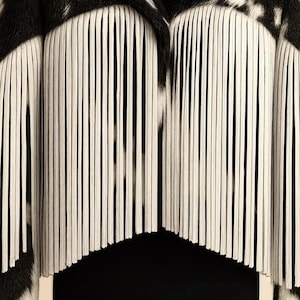 May include: Close-up of a black and white patterned textile with vertical white fringe. The fringe hangs in a curved shape, creating a layered effect. The textile has a zebra-like pattern.