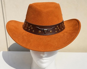 May include: An orange suede cowboy hat featuring a dark brown leather band adorned with metallic studs and a central silver-toned steer skull emblem. The hat has a wide, curved brim and a pinched crown, providing a structured and classic silhouette.