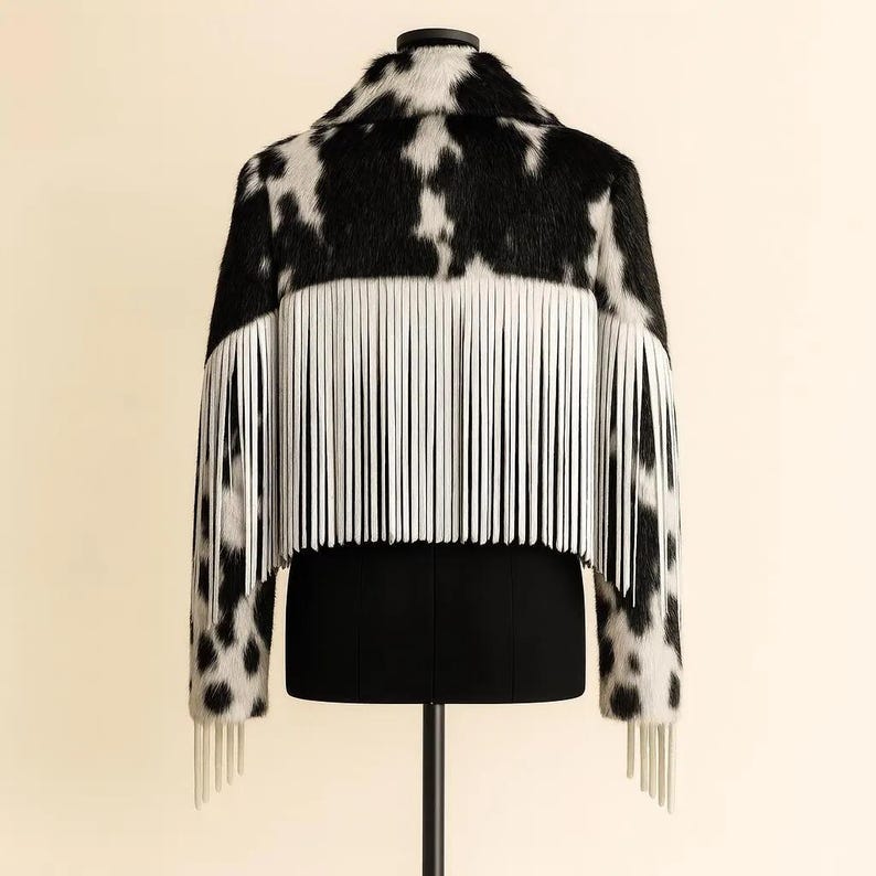 May include: Cropped jacket with a black and white cowhide pattern. The jacket features long sleeves and a collar. White fringe detailing is across the back and on the sleeves. The jacket is displayed on a black mannequin.