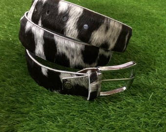 May include: A black and white cowhide belt with a silver buckle. The belt has a natural pattern of black and white patches. The belt is approximately 1.25 inches wide.