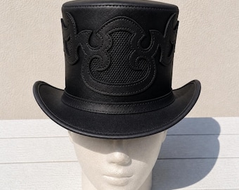 May include: A black leather top hat with an ornate, textured design on the crown. The hat features a wide brim and is displayed on a white mannequin head. The hat's design is symmetrical and detailed.