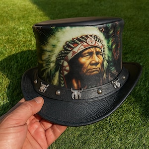 May include: Black leather top hat with a printed image of a Native American chief. The hat features a decorative band with silver-colored bull head accents and rivets. The hat is held against a green grassy background.
