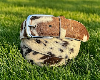 May include: A brown and white cowhide belt with a silver buckle. The belt is coiled on a bed of green grass. The leather has a natural spotted pattern, with shades of brown and white. The buckle is a shiny, rectangular shape.