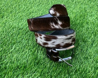 May include: A belt featuring a brown and white cowhide pattern with a textured fur finish. The belt is coiled and includes a silver-toned metal buckle with a prong mechanism, showcasing a distinct spotted appearance across the entire strap surface.