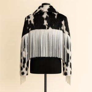 May include: A cropped jacket with a cowhide pattern in black and white, featuring white fringe along the back and sleeves. The jacket is displayed on a black dress form, showcasing its unique design and texture.