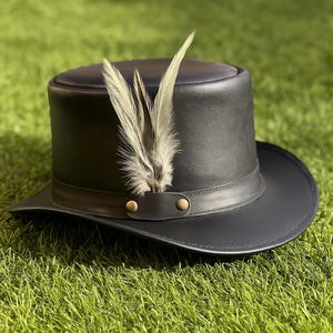 May include: A black leather top hat with a wide brim and a decorative band. The hat features a cluster of light-colored feathers on the side, secured with two brass-colored rivets. The hat is set against a backdrop of green grass.