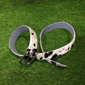 May include: A cowhide belt with a black and white spotted pattern. The belt has a silver-colored buckle and a light blue interior. The belt is displayed on a green surface.
