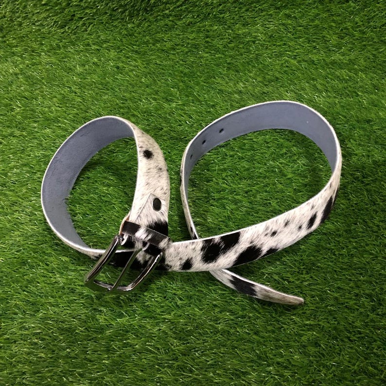 May include: A cowhide belt with a black and white spotted pattern. The belt has a silver-colored buckle and a light blue interior. The belt is displayed on a green surface.