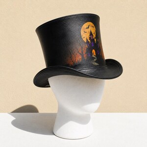 May include: A black top hat featuring a printed graphic of a haunted house, a large yellow full moon, and silhouettes of flying bats. The design includes bare trees and an orange-hued sky, set against a dark, textured background on the hat.