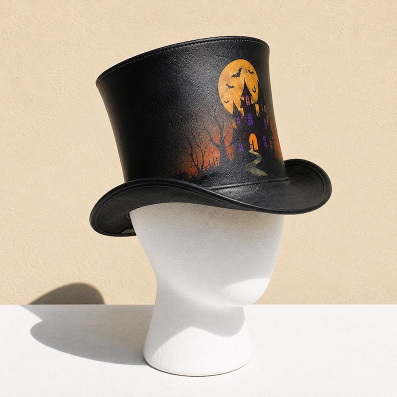 May include: A black top hat featuring a printed graphic of a haunted house, a large yellow full moon, and silhouettes of flying bats. The design includes bare trees and an orange-hued sky, set against a dark, textured background on the hat.
