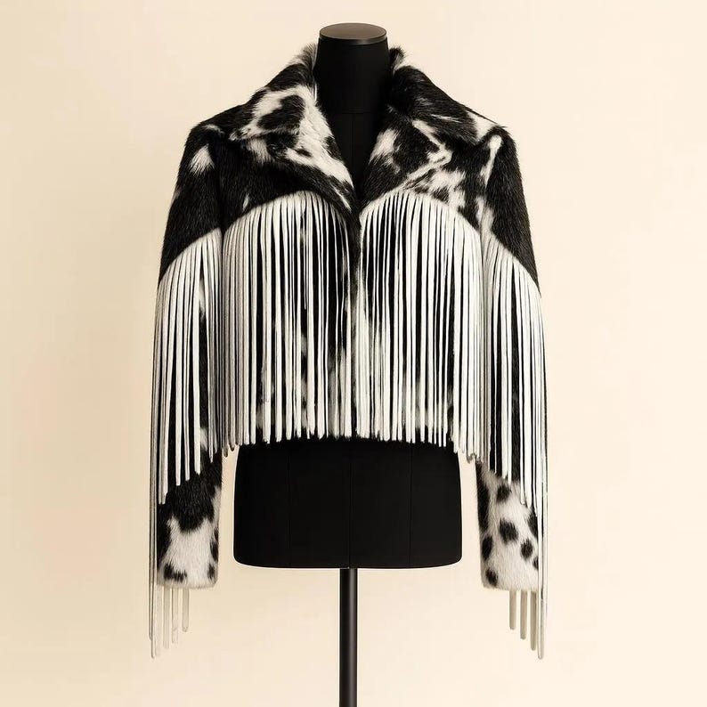 May include: A black and white faux fur jacket with a cowhide pattern. The jacket features long white fringe along the front and sleeves. The jacket is displayed on a black mannequin.