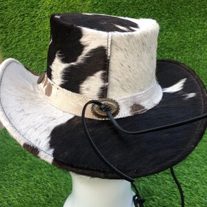 May include: A cowboy hat featuring a cowhide pattern with black and white fur. The hat includes a light-colored band adorned with a metallic oval concho ornament and a thin black leather chin strap that hangs down from the side of the crown.