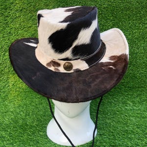 May include: A cowboy hat featuring a cowhide pattern with black, white, and brown patches. The hat has a wide brim, a structured crown, a light-colored band with a circular metallic emblem at the front, and a thin black chin strap cord.
