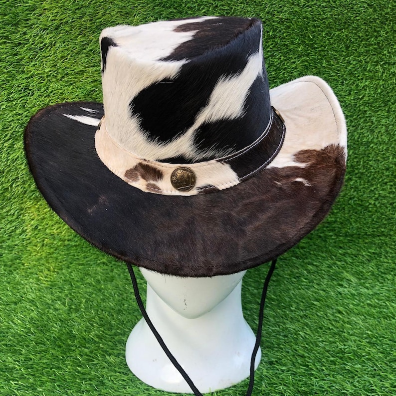 May include: A cowboy hat featuring a cowhide pattern with black, white, and brown patches. The hat has a wide brim, a structured crown, a light-colored band with a circular metallic emblem at the front, and a thin black chin strap cord.