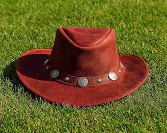 May include: A reddish-brown cowboy hat with a wide brim and a decorative band. The band features silver-colored embellishments. The hat is made of a textured material and sits on green grass.