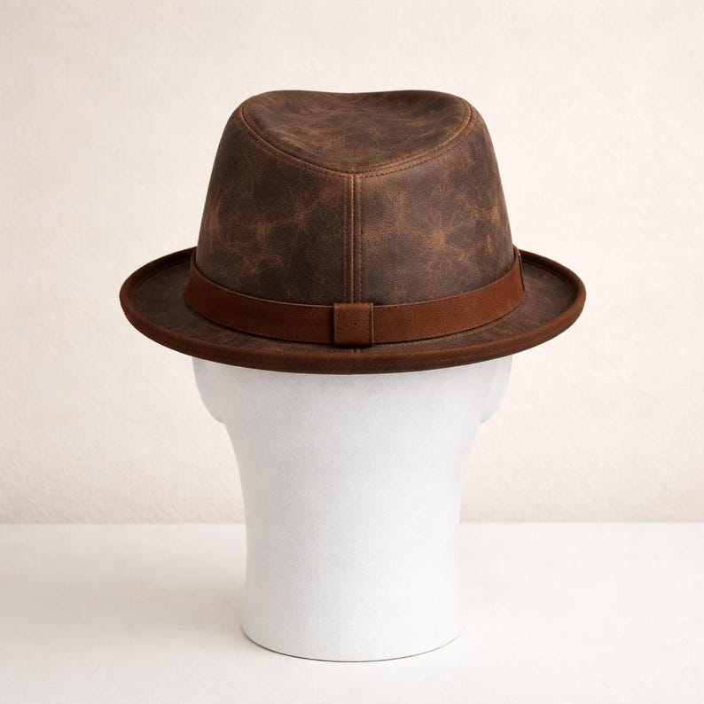 May include: A brown leather fedora hat with a dark brown band. The hat is displayed on a white mannequin head. The hat has a classic fedora shape with a creased crown and a brim that is approximately 2 inches wide.