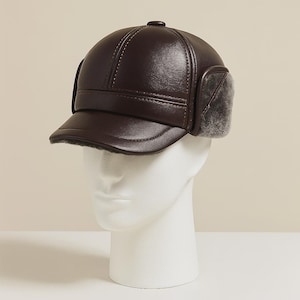 May include: A dark brown leather cap with ear flaps and a short brim. The cap has a rounded crown with stitched details and a small button on top. The ear flaps are lined with a soft, gray fur-like material.