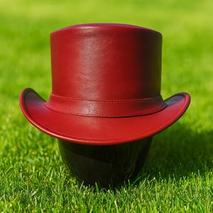 Może przedstawiać: Bordowy skórzany cylinder z szerokim rondem i opaską wokół korony. Kapelusz jest umieszczony na zielonej trawie, podkreślając jego formalny design i bogaty kolor. Ten klasyczny kapelusz jest odpowiedni na specjalne okazje.