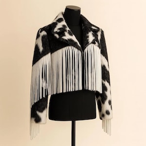 May include: A black and white patterned jacket with white fringe detail. The jacket is displayed on a black mannequin. The pattern resembles a cowhide print. The fringe is along the front and sleeves of the jacket.