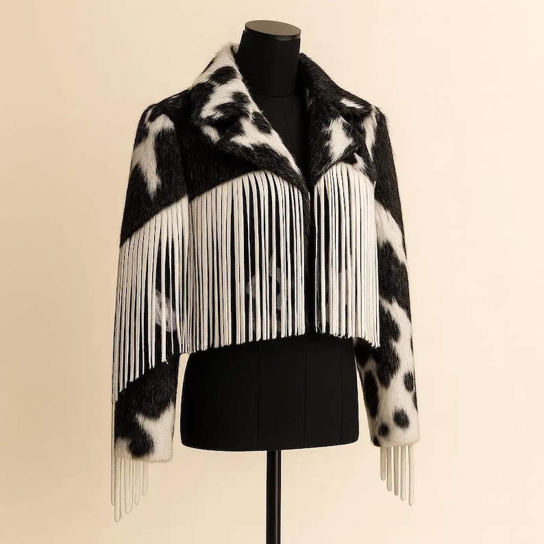 May include: A black and white patterned jacket with white fringe detail. The jacket is displayed on a black mannequin. The pattern resembles a cowhide print. The fringe is along the front and sleeves of the jacket.