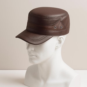 May include: A brown leather military-style cap with a short brim. The cap has a structured crown and a decorative band around the middle. Two small metal eyelets are visible on the side of the cap. The cap is displayed on a white mannequin head.