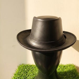 Peut inclure: Un chapeau fedora noir avec une finition lisse et mate et une calotte structurée. Le chapeau présente un bord large et plat avec un détail de bordure tissé et texturé, ainsi qu'une bande noire assortie entourant la base de la calotte.